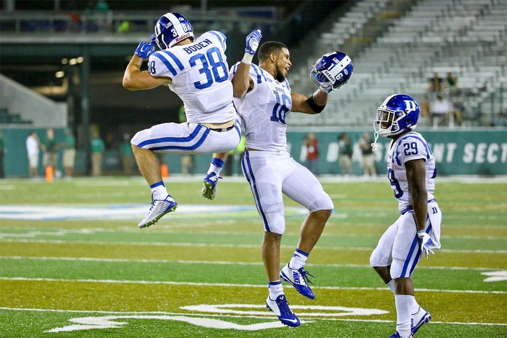 Three football players on the field celebrating. Two are jumping in the air as one stands nearby